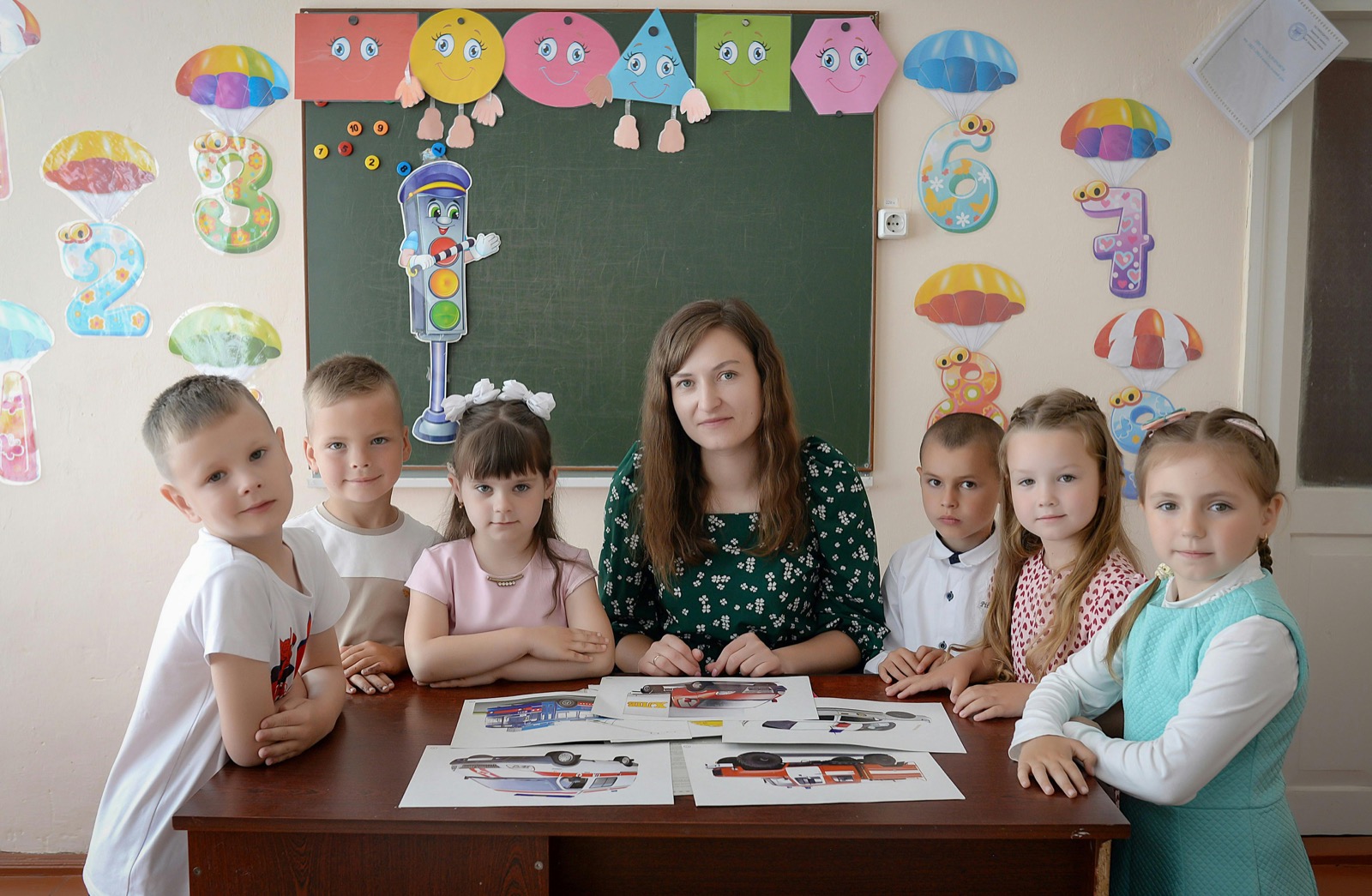 Teacher and children gathered around a classroom table in a colorful room.