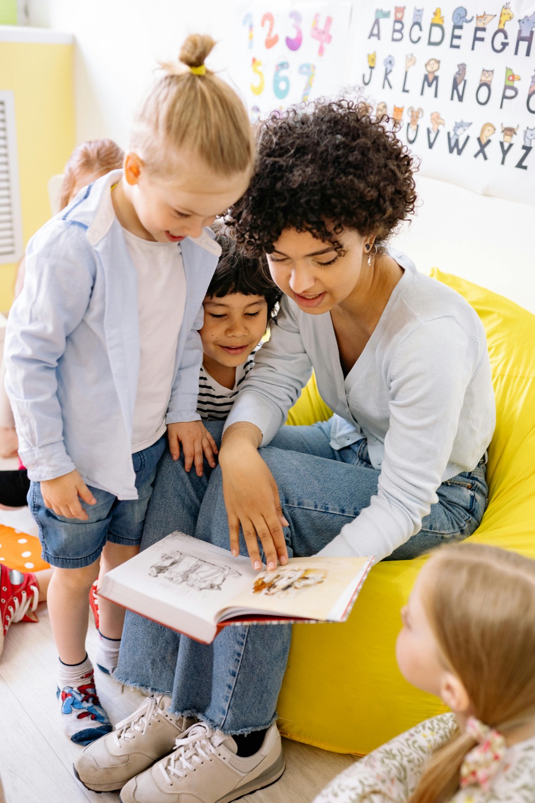 Teacher reading with young students gathered nearby in a bright classroom.